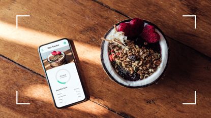Sunlight coming through window onto wooden table featuring phone with calorie counter app open and nutritious fruit breakfast in a bowl, representing how many calories should I eat in a day