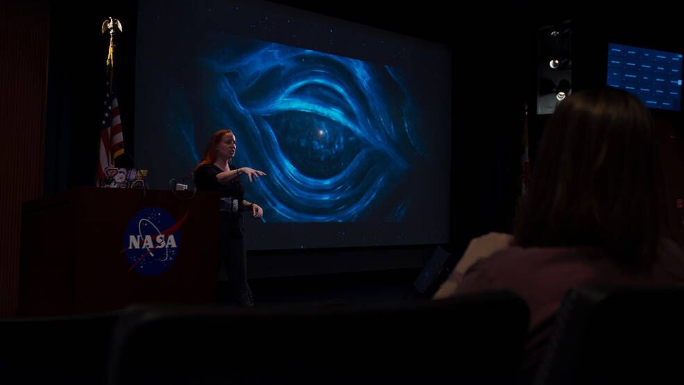 a woman gives a lecture in a dark room with space imagery projected on a screen in the background.