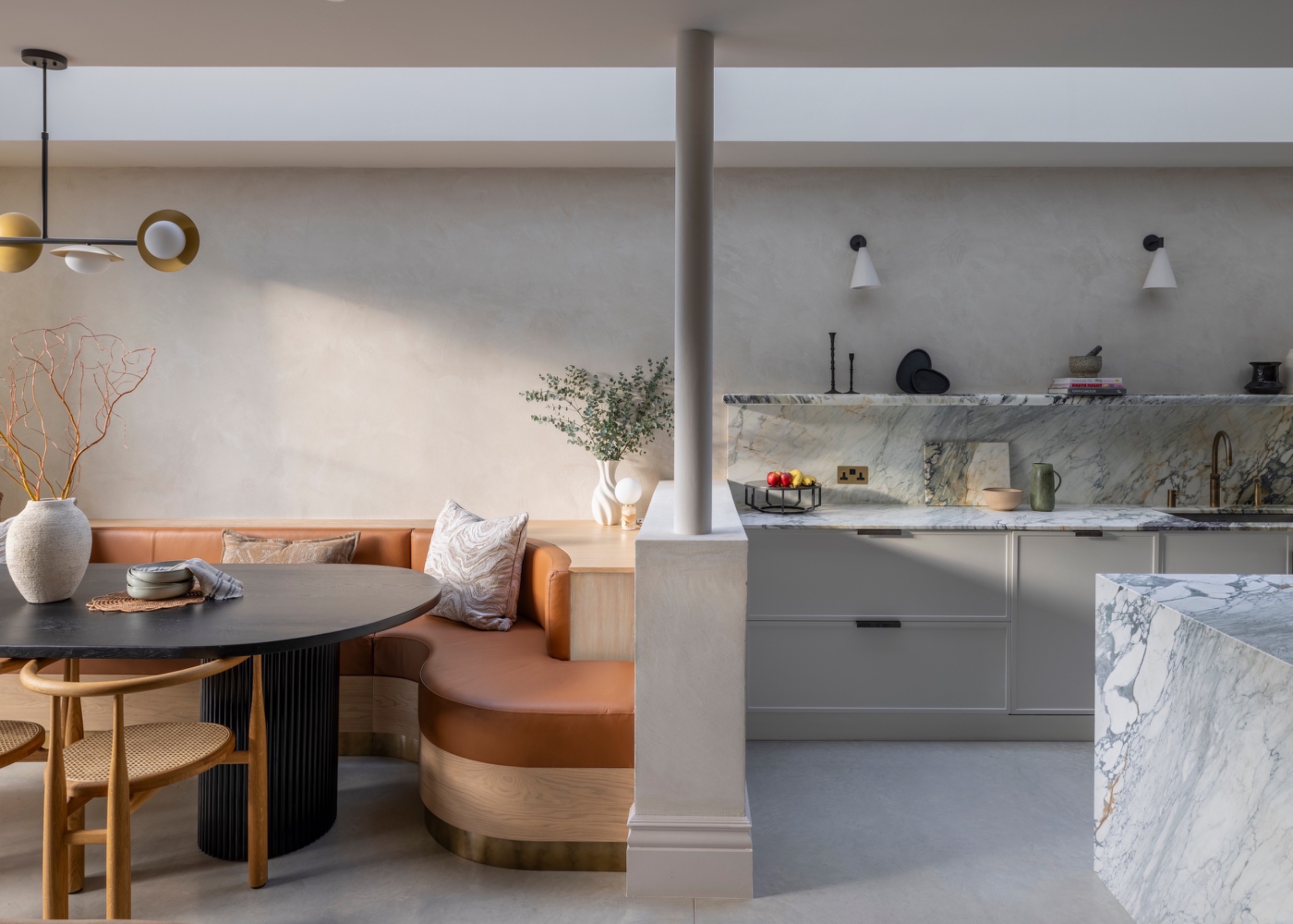 Low partition incorporating a structural column separates a curved tan leather banquette and dark pedestal dining table from pale kitchen cabinetry with veined porcelain countertops.