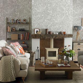 Living room with light brown and white woodland wallpaper, a white woodburner fireplace, cosy white sofa and large wooden square coffee table