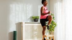 woman in exercise clothes a pink vest and black cycling shorts stepping onto a small indoor step and raising her knee. there's a wooden unit, net curtains and a blank wall behind her.