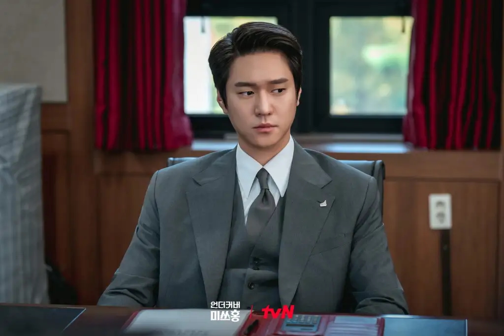 A man wearing a suit (Go Kyung-pyo) sits in an office conference room, in the Netflix K-drama 'Undercover Miss Hong.'
