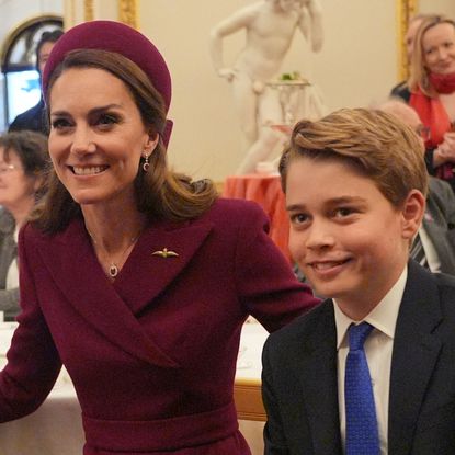 Kate Middleton wears a burgundy coat and matching hat and poses with son Prince George, who is wearing a navy suit, white shirt, and blue tie