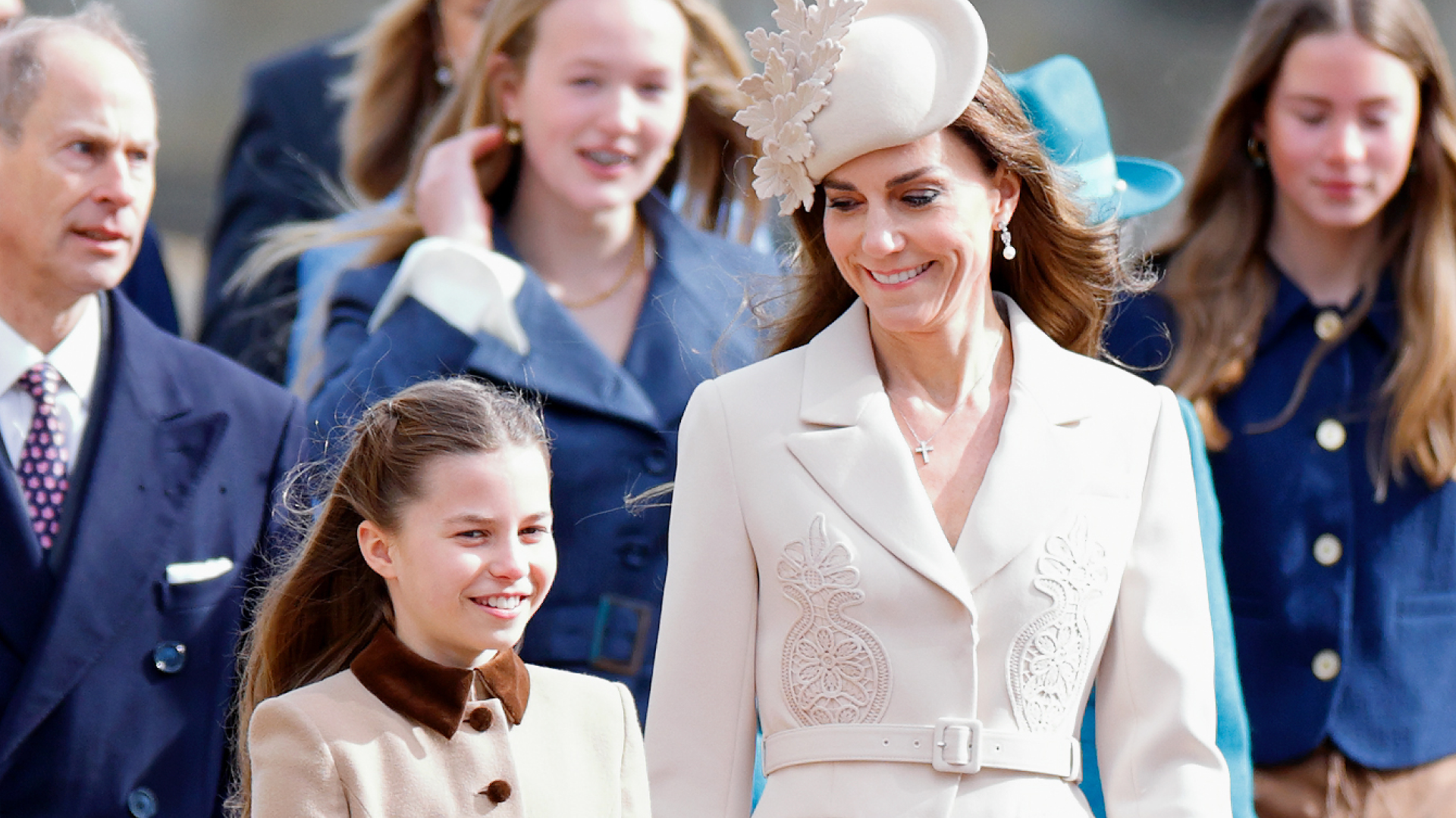 Princess Charlotte wears a tan brown coat dress and walks next to mom Kate Middleton who wears a cream ensemble