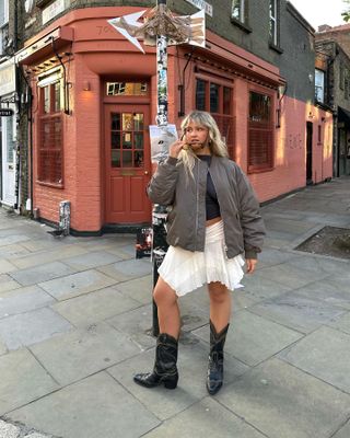model wears white flowy miniskirt, black top, bomber jacket, and black cowboy boots in front of a brick-red building