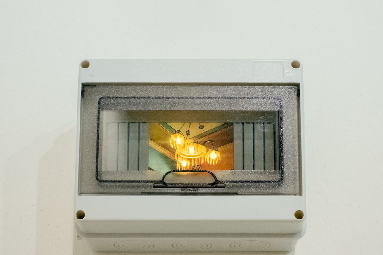 A chandelier glowing with warm light is captured in a photograph back-lit inside an electricity box as part of an art installation.