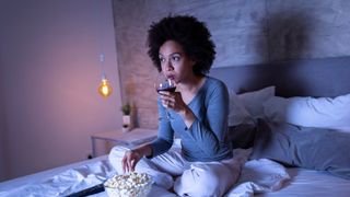 A woman sat crossed legged in bed eating popcorn and drinking wine