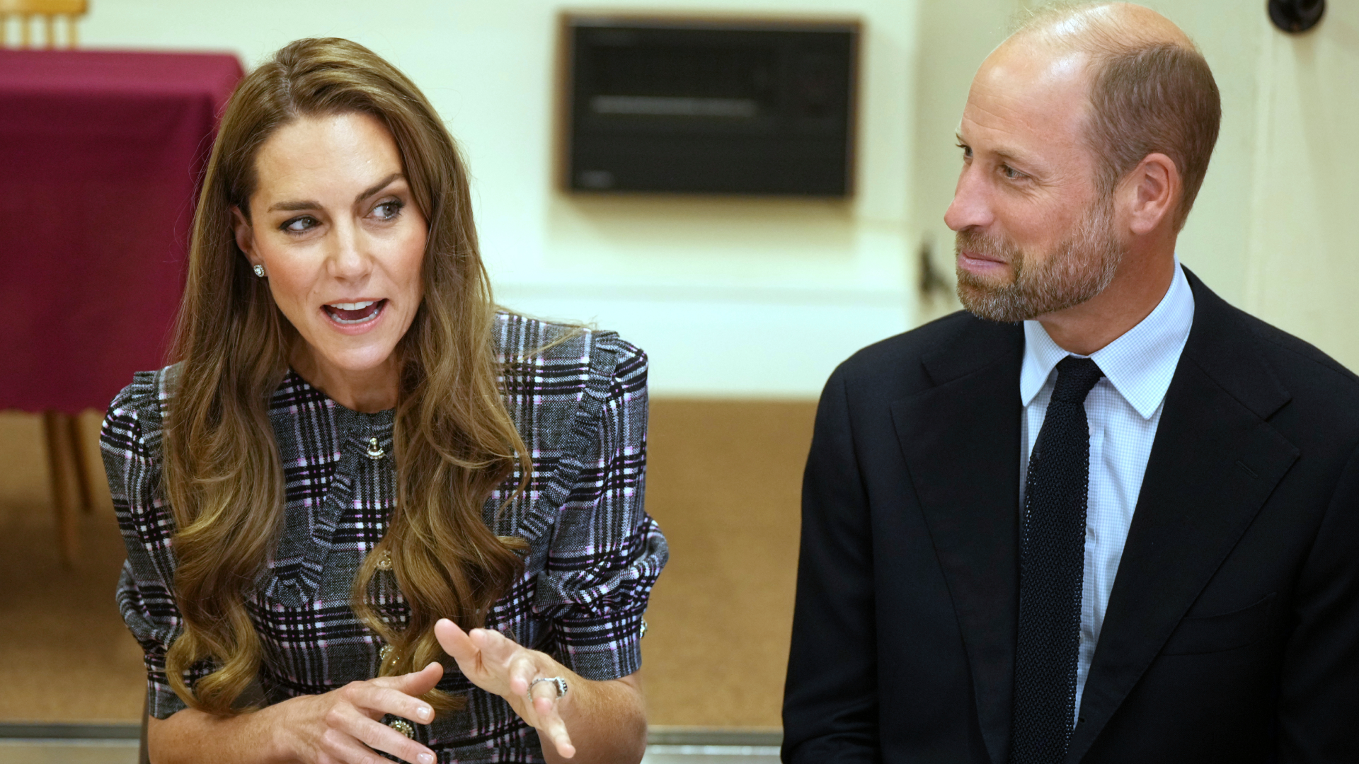 Prince William wearing a dark suit sitting next to Kate Middleton and looking at her, smiling, as she talks and gestures with her hands
