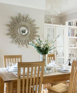 A dining room with a gray wall with a silver sun-shaped mirror, a light wooden dining table set laid out with white and glass dinnerware and a vase of green flower stems and a white bookshelf