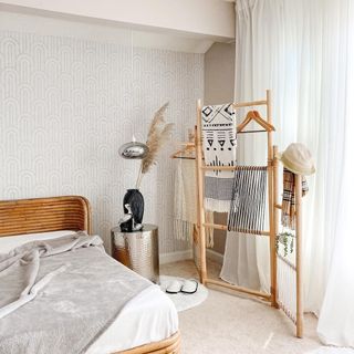 Bamboo room divider hung with mono patterned throws and clothes in neutral bedroom with subtle geo wallpaper, and natural textures.