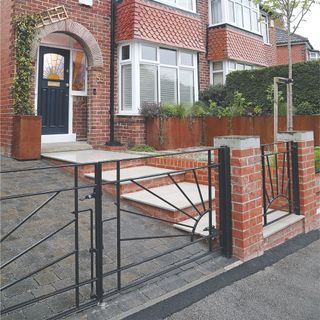 Black metal gate framing sloped driveway with block paving next to steps leading to brick house