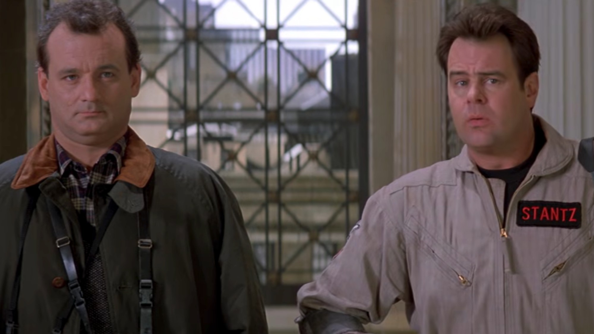 Bill Murray in a green coat with a brown collar, standing next to Dan Aykroyd in his Ghostbuster uniform as they walk into the museum in Ghostbusters II. 