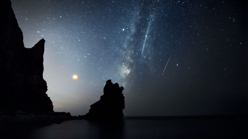 Two meteors are pictured streaking through a star-studded sky alongside the glowing band of the Miilky Way towards a dark ocean. Rocky outcrops are visible to the left of the screen, where a subtle glow creeps skyward.