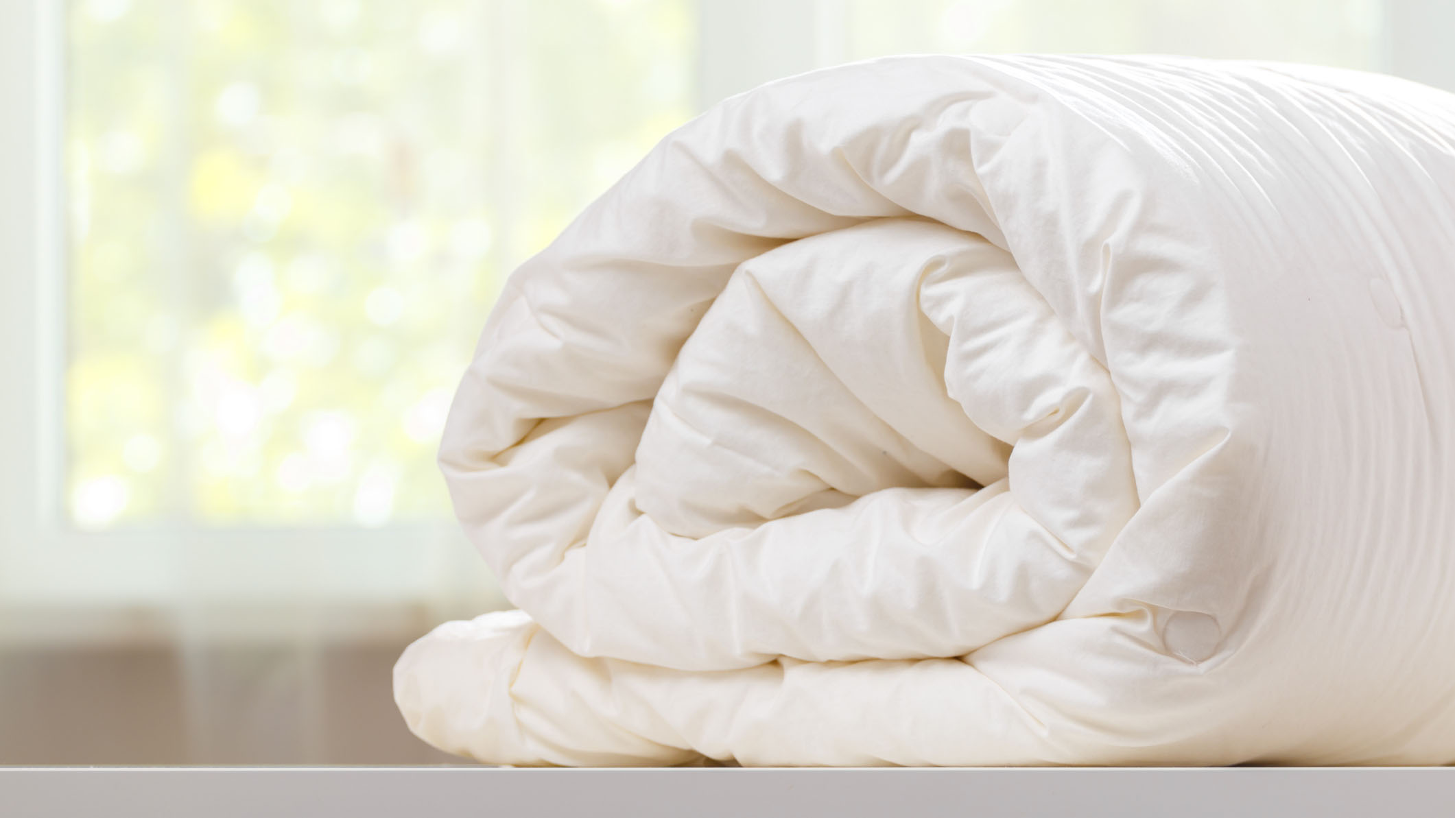 Как заправить кровать по скандинавскому методу сна. На изображении показано свернутое белое пуховое одеяло и положенное на белую столешницу.