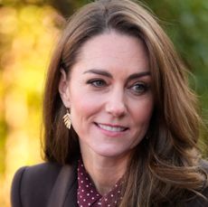 Kate Middleton wearing a brown coat, gold fern earrings and burgundy dress smiling as she looks to the side