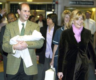 Prince Edward wearing a checked green blazer holding newborn Lady Louise Windsor and smiling as he leaves the hospital with Duchess Sophie, wearing a brown coat and pink scarf and smiling