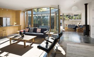 A sitting area with a navy sofa and chairs set around a table, and a kitchen in light wood. A planted, glassed-off circular garden can be seen across from the open concept space.