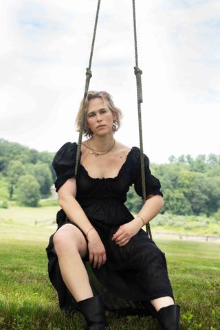 tommy dorfman wears a black dress black boots and several necklaces while sitting on a swing in a field