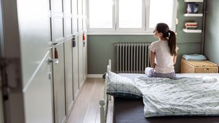 A brunette woman with a ponytail sitting on the bed of her childhood bedroom.