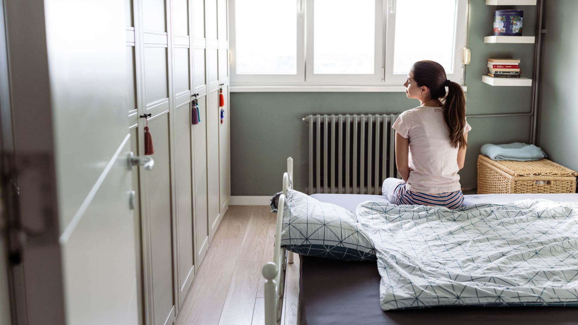 A brunette woman with a ponytail sitting on the bed of her childhood bedroom.