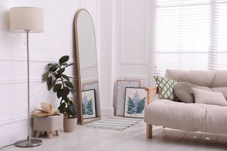 A modern white room, decorated with a large wooden arch mirror, a plant, a cream sofa and other framed photos.