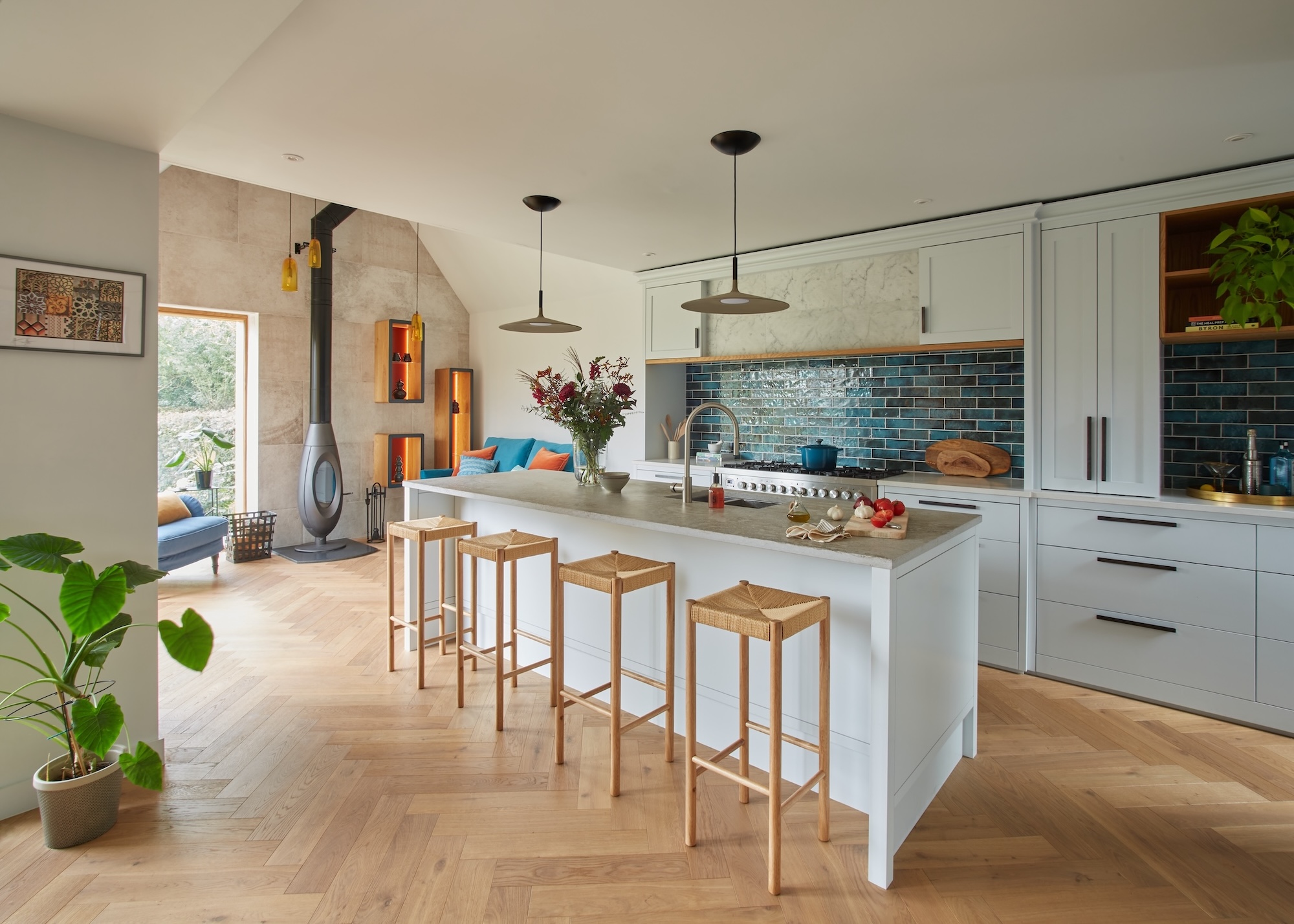 A modern kitchen with a small kitchen island and two pendant lights above, the floors are wood herringbone with a wood burner on the far wall.
