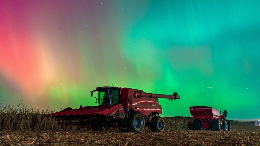red, magenta and green auroras appear as curtains of light in the sky above farming equipment in a field.