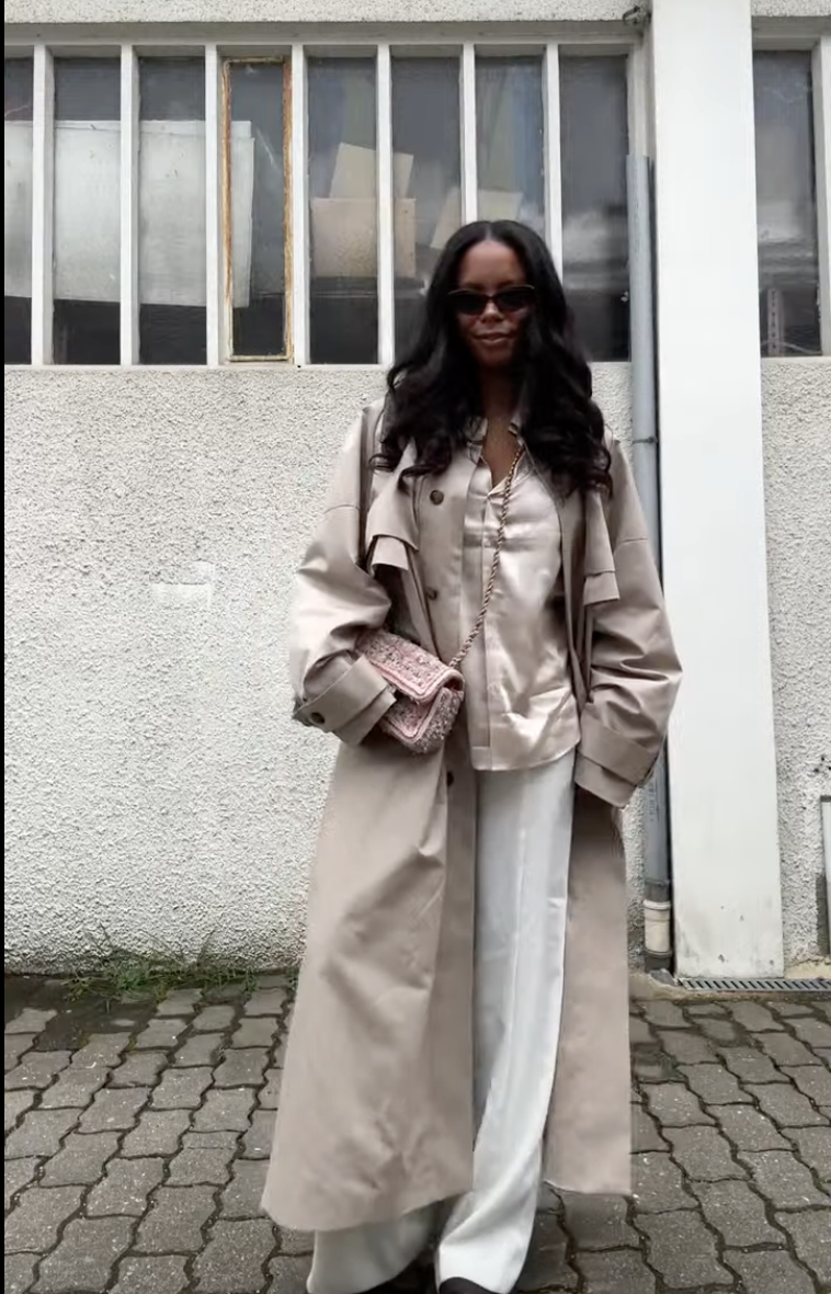 Woman wearing a satin shirt, white trousers, and a long tan trench coat.