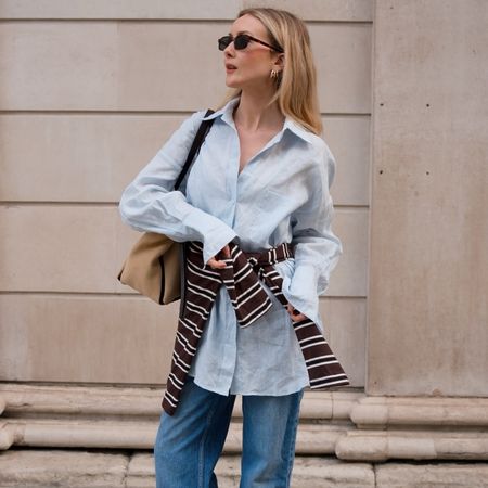 British style influencer Chloe Butler poses on a London sidewalk wearing sunglasses, an oversize light-blue linen button-down shirt, a striped sweater around the waist, tan bag, and jeans