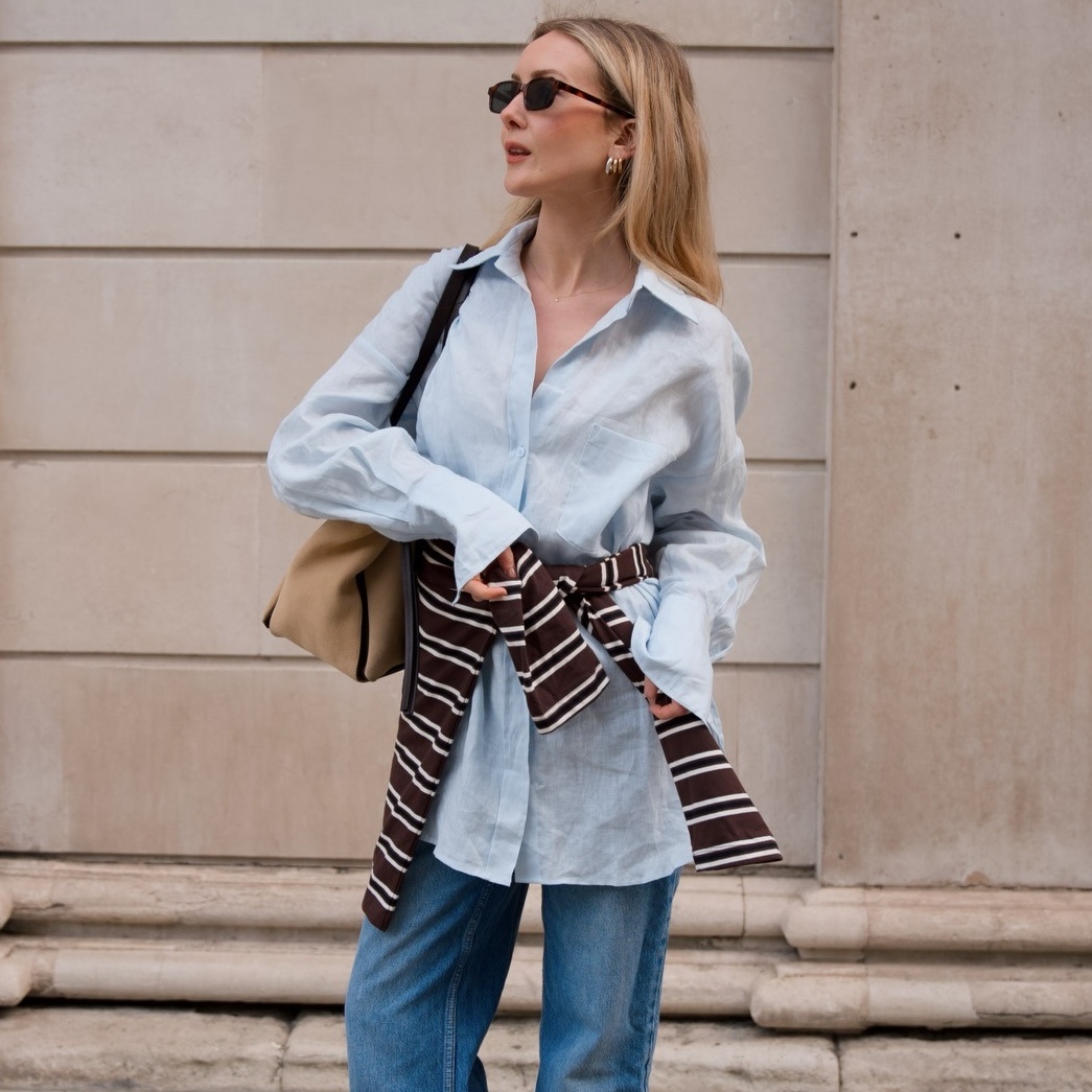British style influencer Chloe Butler poses on a London sidewalk wearing sunglasses, an oversize light-blue linen button-down shirt, a striped sweater around the waist, tan bag, and jeans