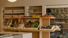 A sideway view of a luxury supermarket with wooden open shelving filled with fruit and vegetables, jars of pickled products behind them, and bags of vegetables sitting in an open fridge.