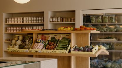 A sideway view of a luxury supermarket with wooden open shelving filled with fruit and vegetables, jars of pickled products behind them, and bags of vegetables sitting in an open fridge.
