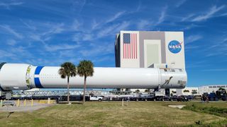 a large white rocket rolls down a road with a big nasa building and blue skies in the background.