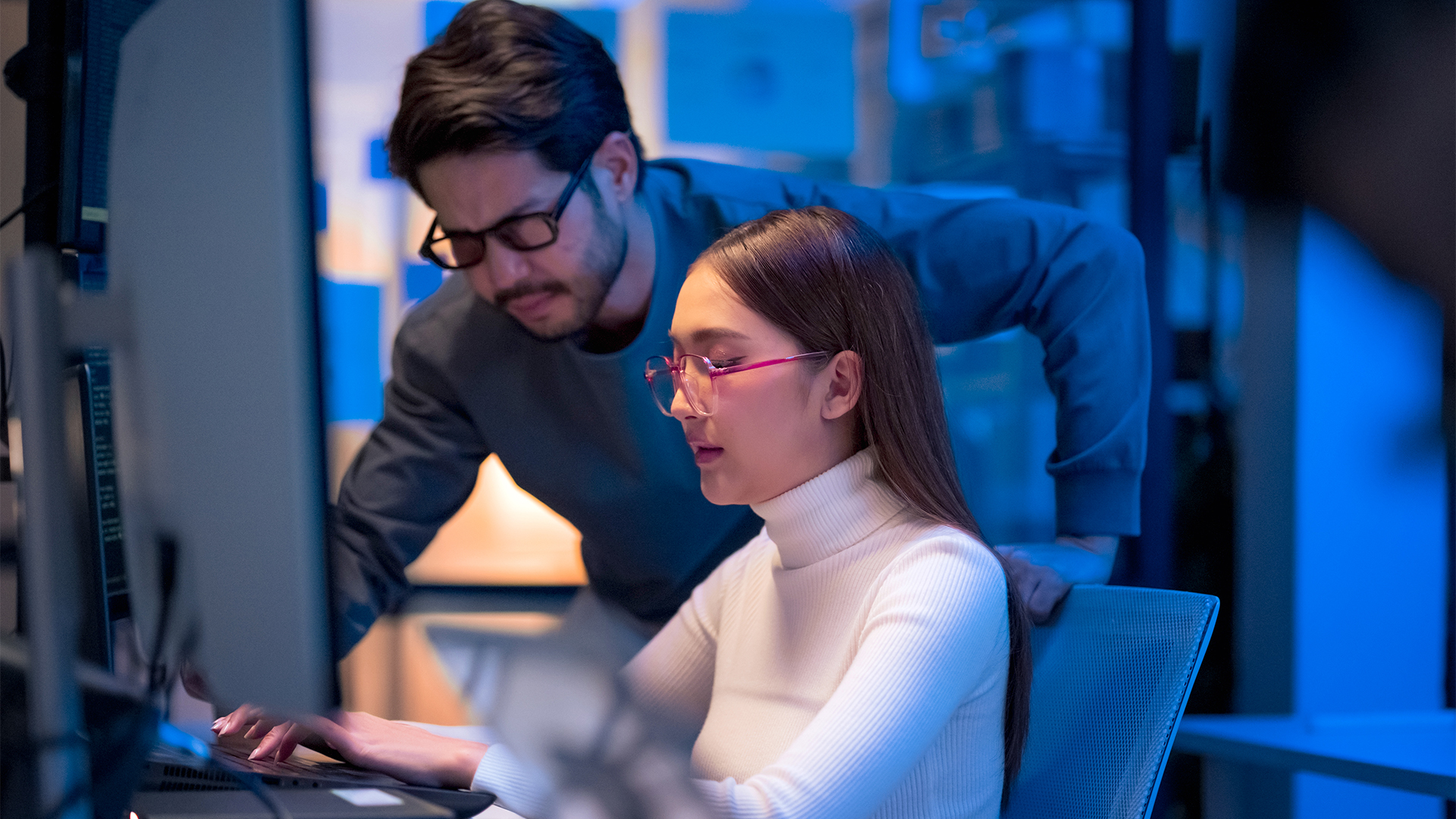 Female office worker working at desktop computer station with colleague leaning over shoulder and watching screen activity. 