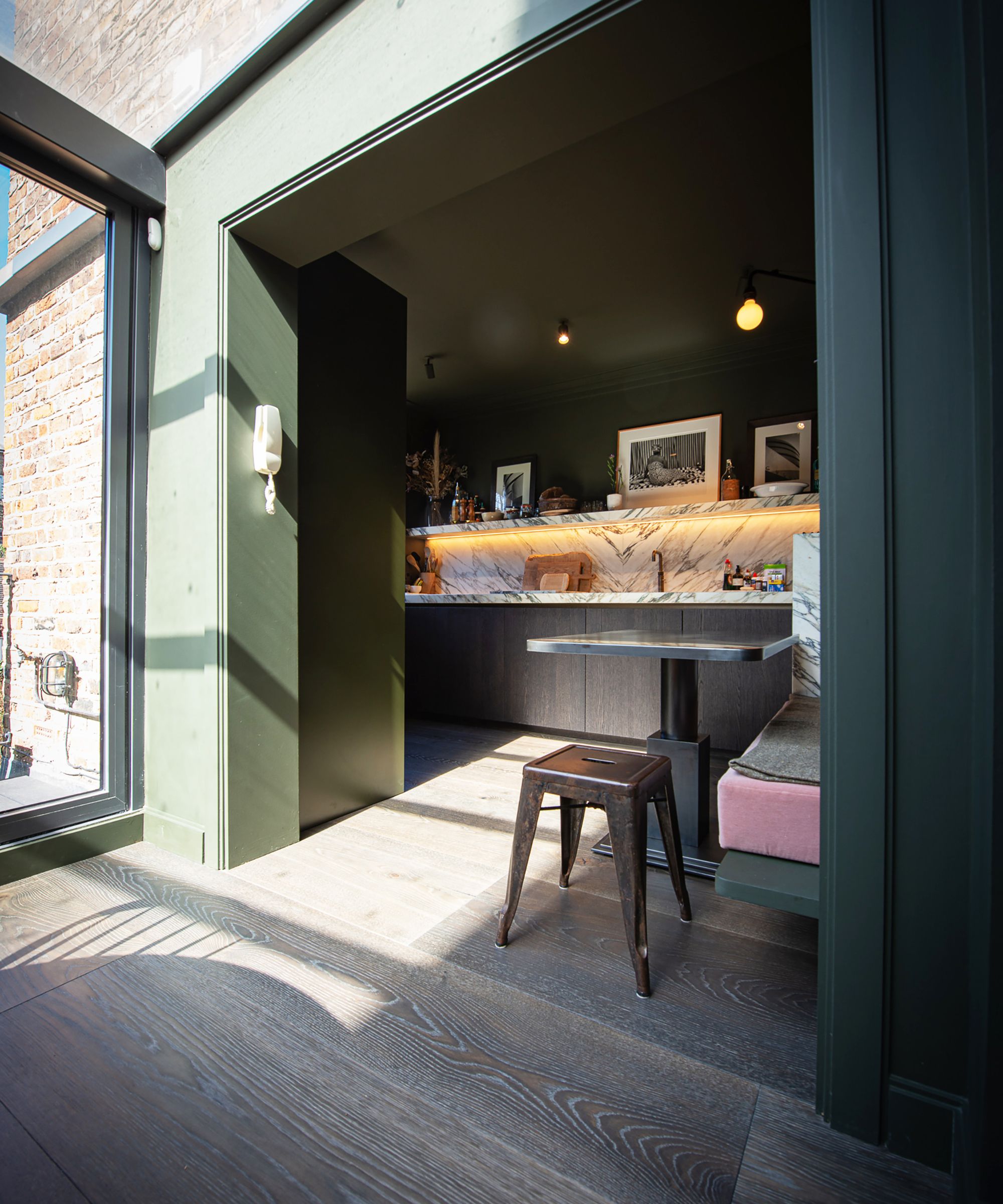 A dark green painted kitchen alcove with a dramatic veined marble worktop and splashback, dark oak cabinetry, industrial metal stool, bare bulb pendants and wide plank oak flooring.