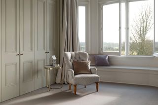 Neutral upholstered chair in carpeted bedroom space with light green built-in wardrobe doors, a seated bay window and gilded side table.