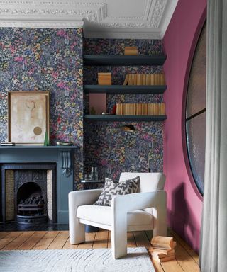 A living area with a blue floral wall with navy blue wall shelves, a deep blue fireplace, a white modern angular armchair, a light wooden floor with a gray fuzzy rug, and a burgundy wall
