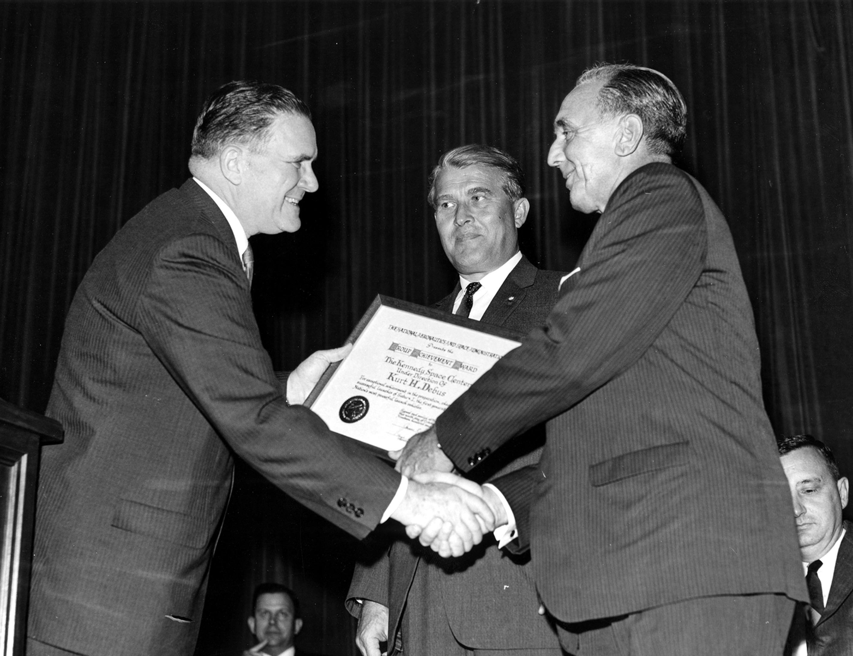 Two men shaking hands and smiling, a third stands in the background.