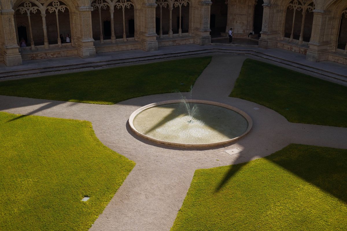 Circular fountain surrounded by geometric lawns and cloister arches, partly illuminated by strong sunlight.