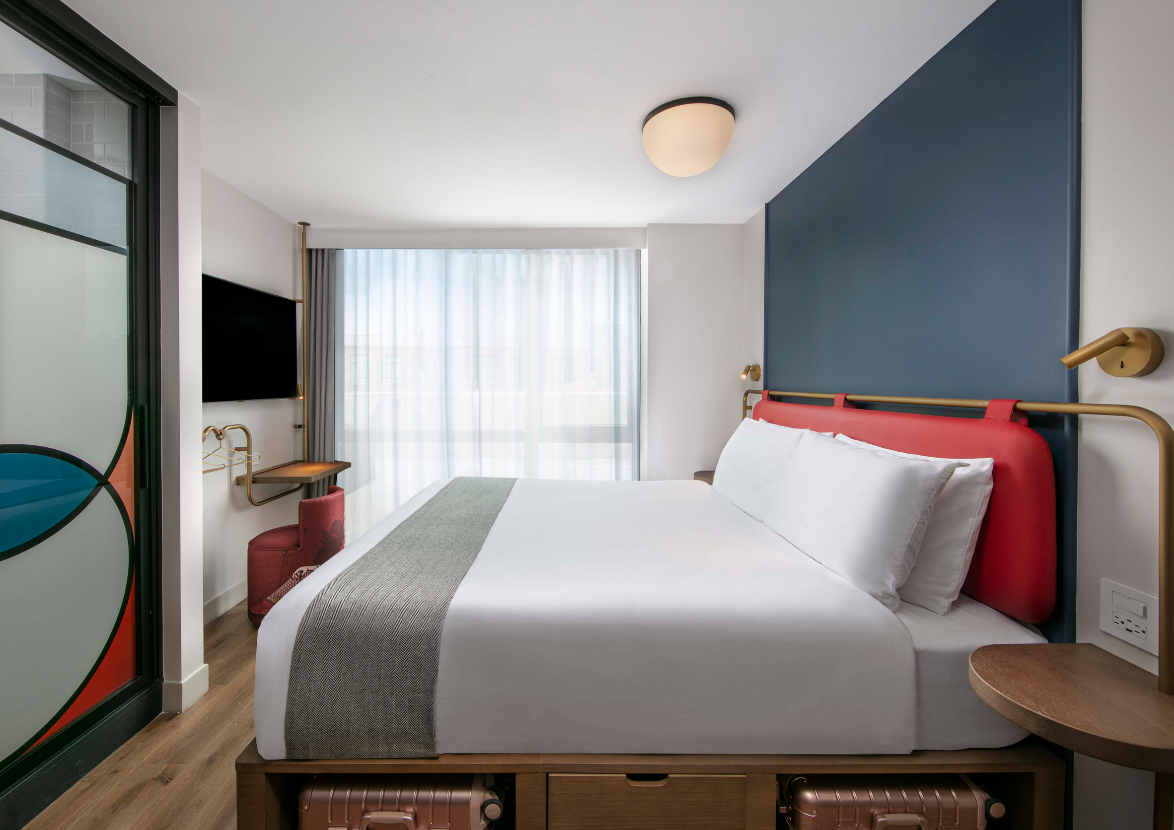 A Bauhaus-inspired hotel room features a quirky color palette of muted blues, reds, oranges, and creams, manifesting itself across soft-angled furniture with a modernist style.