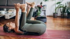 side shot of a woman in khaki leggings on a red exercise mat and wooden floor in a happy baby yoga pose, on her back knees close to her chest and holding her feet. there's a blurred living room setting behind her.