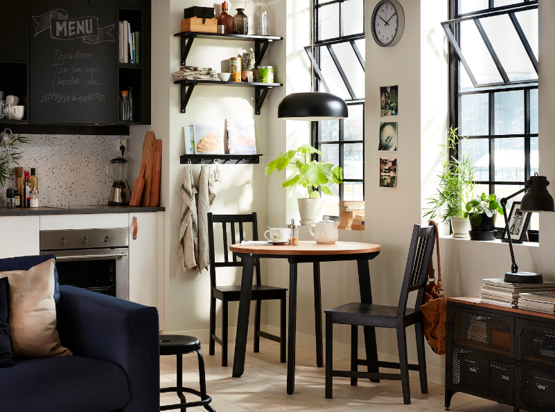 a small loft apartment with dark furniture, including a sofa, a wooden dining table, two windows with dark frames and a dark kitchen to the left