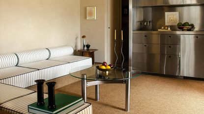 Image of a beige living room with a chrome home bar built into the wall. There is a low sectional bench seat with bolster pillows and a striped pattern.
