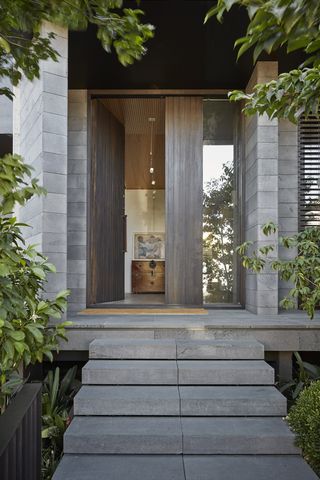 A residential home built with shades of grey concrete with a full tall front door which is open. Grey concrete stair cases leading to the open front door of a grey concrete residential home. The front door is a tall wood design with a beige door carpet