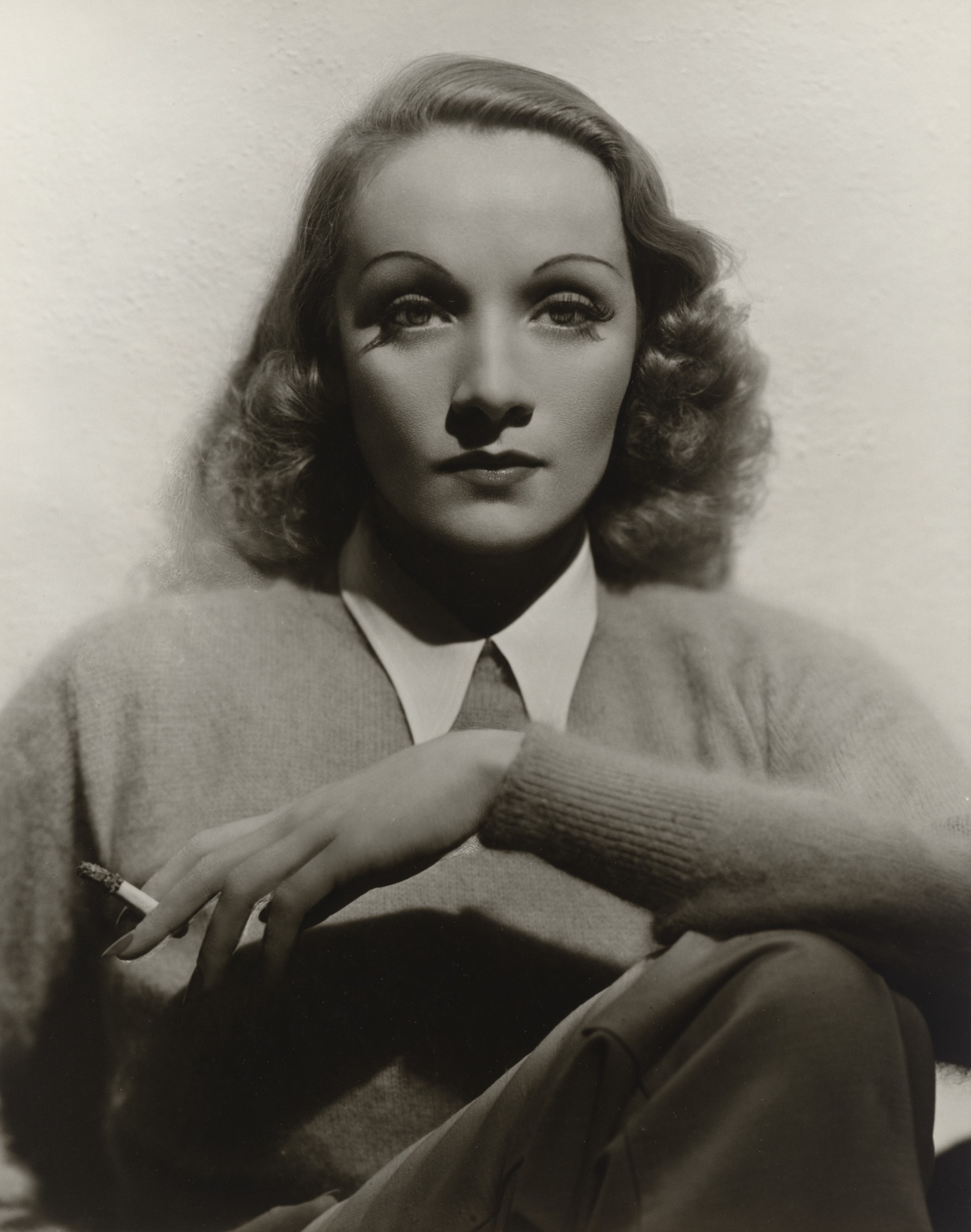 Marlene Dietrich, lit from above, sits with her arm resting on her knee and a cigarette between her fingers, looking straight at the camera with heavy-lidded eyes.