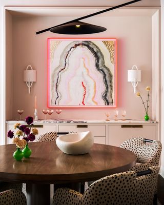 Image of a light pink dining room with a round wooden dining table and four cheetah print chairs around it. There is an abstract painting on the back wall with a neon pink boarder and two white wall sconces on either side.