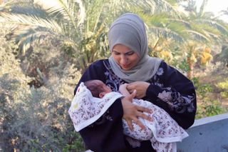 Woman wearing a hijab holding her baby with palm trees in the background