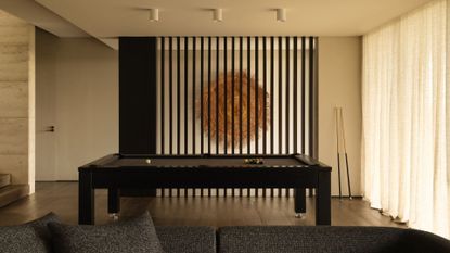 A white room with dark wood paneling, a black pool table in the middle with cues leaning on the wall, and a gray couch in front