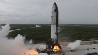 a tall pointy chrome rocket with black belly and white frosty mid section fires its engines on a test stand with smoke billowing to the left.