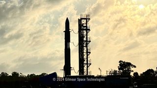 the silhouette of a rocket stands next to a launch tower with staircase, against a bright cloudy sky.